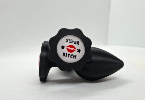Czarny korek analny „Super Bitch” – silikonowy butt plug z białym napisem, czerwonym pocałunkiem i gwiazdkami.